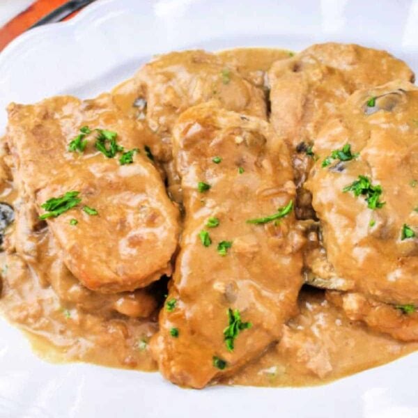 Four pork chops covered in creamy brown mushroom gravy, garnished with chopped parsley on a white plate—a classic Slow Cooker Pork Chops and Gravy dish.